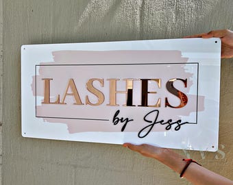 May include: A white sign featuring the text "LASHES by Jess" in rose gold and black. The sign has a pink watercolour-style background and a black rectangular border. The sign is held up by two hands.