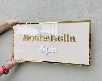 May include: A gold framed sign with a blush pink background. The sign says "Mochabella Spa" in gold and white lettering.