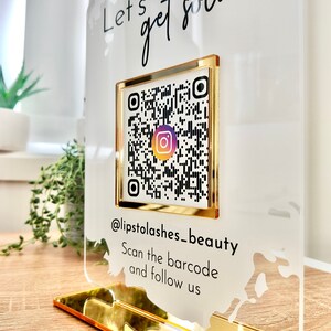 Lets get social sign with white splash background and gold mirror combination, QR code in the middle with Instagram logo, and instagram username under the QR code, and instruction to scan the Qr code to follow under the handle