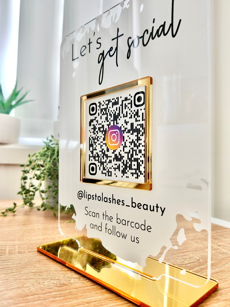 Lets get social sign with white splash background and gold mirror combination, QR code in the middle with Instagram logo, and instagram username under the QR code, and instruction to scan the Qr code to follow under the handle