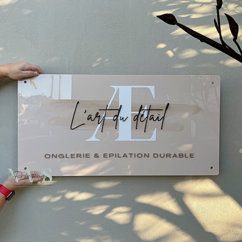 custom rectangle acrylic business sign wall, beige blush pink acrylic backing with logo on it, AE printed in white, business name in 3D black acrylic across, business tagline in 3D taupe acrylic along the bottom with 4 mounting holes on each corners