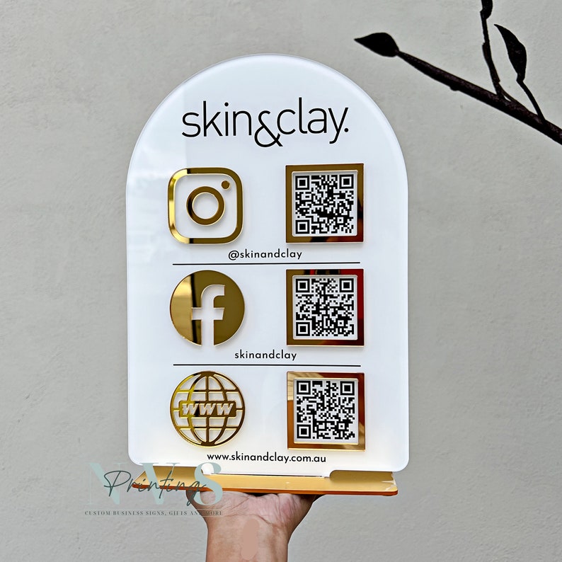 arch QR Code social media display stand in white acrylic, skin business logo on top, 3D instagram facebook website logo in gold mirror acrylic along with each QR codes on the left, instagram facebook handle and website address written under QR code
