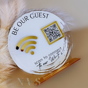 Op de afbeelding: Rond wit acryl bord met gouden accenten. Het bord zegt "Be Our Guest" en heeft een wifi-symbool en een QR-code. De tekst onder de QR-code zegt "Scan to connect to our Wi-Fi".