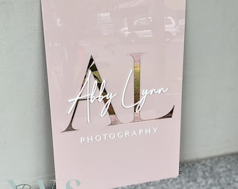 May include: Pink acrylic sign featuring the letters "AL" in a white outline with a mirrored finish. The letters are cut out to reveal a floral pattern. The text "Abby Lynn Photography" is written in white cursive below the letters.