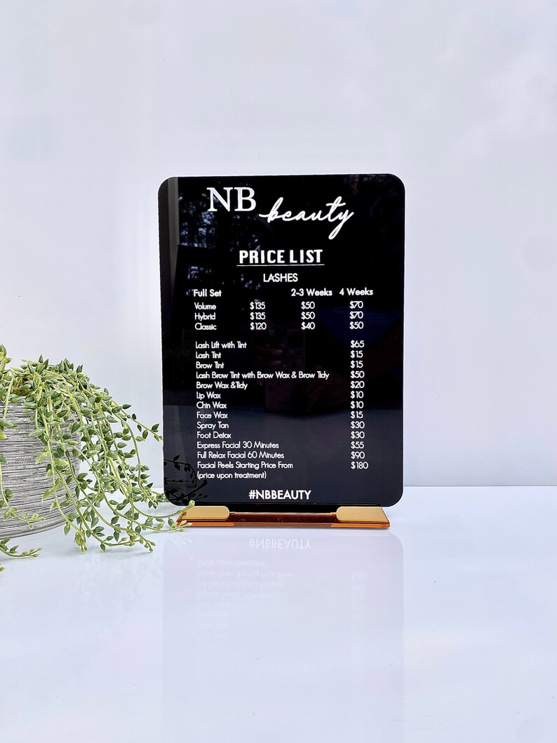 May include: Black acrylic sign with white text listing prices for beauty services, including lashes, waxing, and facials. The sign reads "NB beauty" and "#NBBEAUTY".