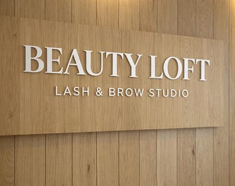 Può includere: Un'insegna in legno con la scritta "BEAUTY LOFT" in grandi lettere bianche. Sotto, in lettere bianche più piccole, si legge "LASH & BROW STUDIO". L'insegna è montata su una parete di legno chiaro a pannelli verticali.