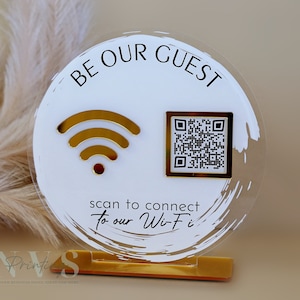 Op de afbeelding: Een rond wit acryl bord met gouden accenten. Het bord zegt "Be Our Guest" en heeft een wifi-symbool en een QR-code. Het bord staat op een gouden standaard.