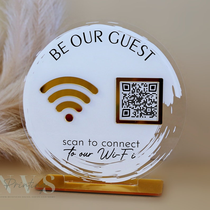 Op de afbeelding: Een rond wit acryl bord met gouden accenten. Het bord zegt "Be Our Guest" en heeft een wifi-symbool en een QR-code. Het bord staat op een gouden standaard.