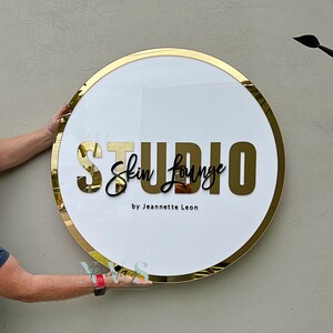 Acrylic Round Business Sign against the wall in white acrylic backing with gold mirror round border frame and 3D letters in gold mirror acrylic and black acrylic, skin lounge studio in 3D on the middle and business owner as business tagline