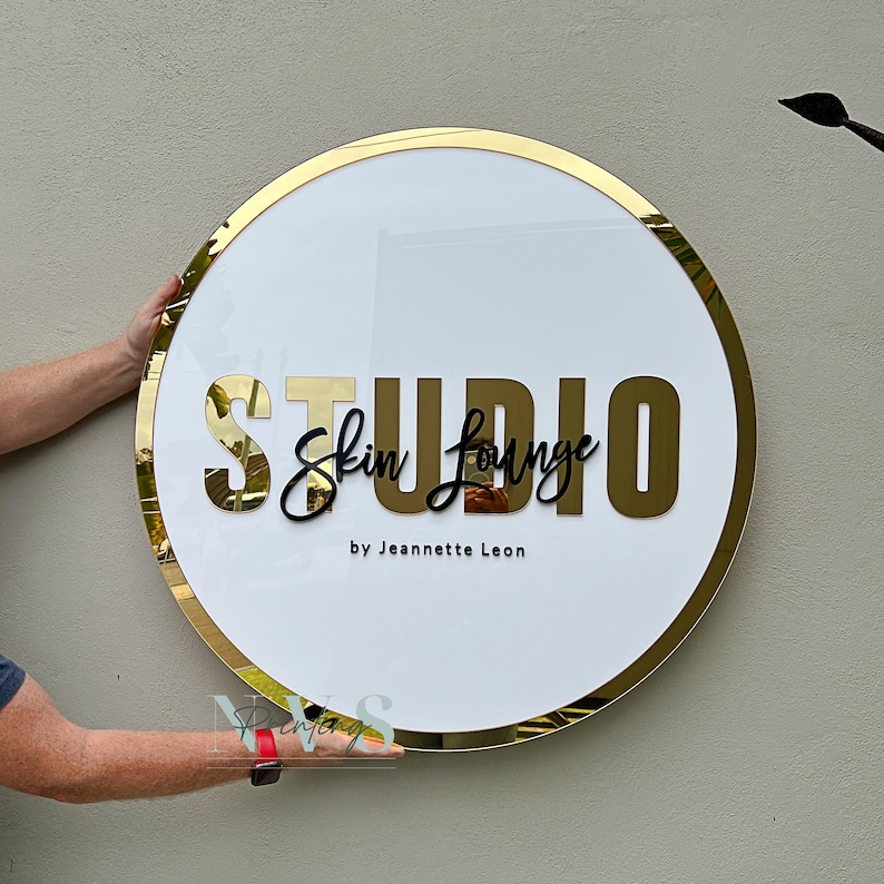 Acrylic Round Business Sign against the wall in white acrylic backing with gold mirror round border frame and 3D letters in gold mirror acrylic and black acrylic, skin lounge studio in 3D on the middle and business owner as business tagline
