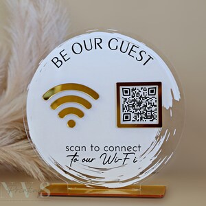 Op de afbeelding: Een rond, wit acryl bord met gouden accenten. Het bord zegt "Be Our Guest" en heeft een gouden wifi-symbool en een zwart-witte QR-code. De tekst onder de symbolen zegt "scan to connect to our Wi-Fi".