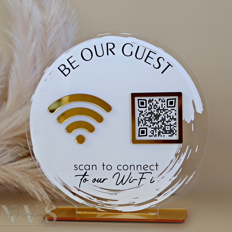 Op de afbeelding: Een rond, wit acryl bord met gouden accenten. Het bord zegt "Be Our Guest" en heeft een gouden wifi-symbool en een zwart-witte QR-code. De tekst onder de symbolen zegt "scan to connect to our Wi-Fi".