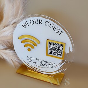 Op de afbeelding: Een rond wit acryl bord met gouden accenten waarop "Be Our Guest" staat en een wifi-symbool en een QR-code. Het bord staat op een gouden standaard. Scan om verbinding te maken met onze wifi.