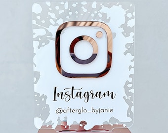 May include: A white acrylic sign with a rose gold Instagram logo and the text "Instagram @afterglo_byjanie". The sign has a white splatter paint design and is on a rose gold stand.