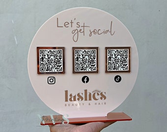 May include: A white circular sign with the text "Let's get social" in rose gold lettering. Three square QR codes are displayed below the text, each with a corresponding social media icon: Instagram, Facebook, and TikTok. The sign is on a rose gold stand and reads "lashes BEAUTY & HAIR".