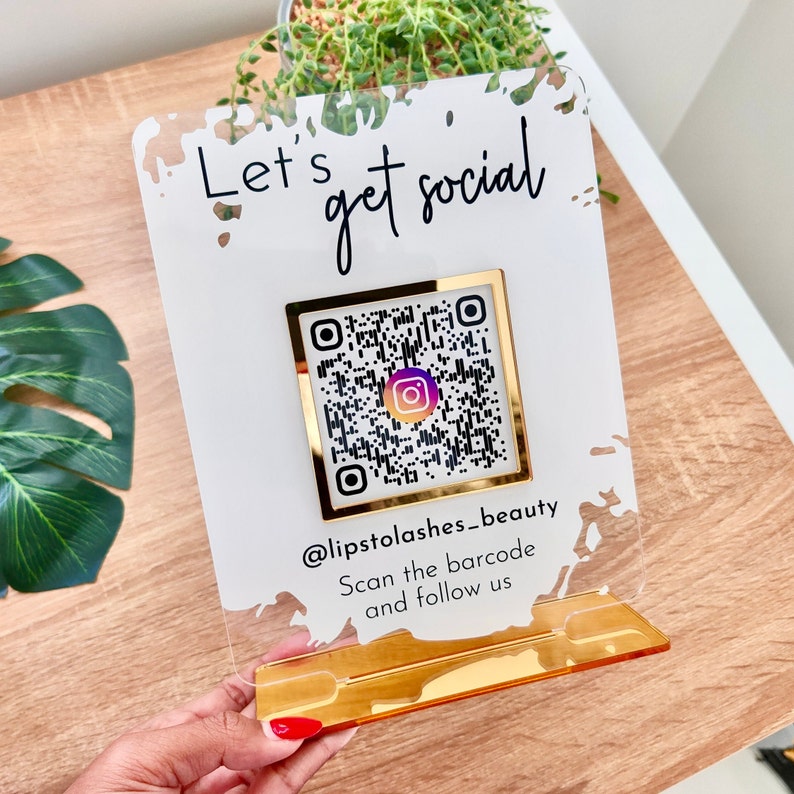 Lets get social sign with white splash background and gold mirror combination, QR code in the middle with Instagram logo, and instagram username under the QR code, and instruction to scan the Qr code to follow under the handle