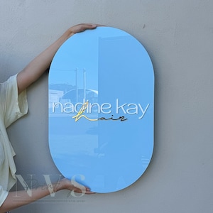 May include: A light blue, oval-shaped sign with the words "nadine kay hair" in white and gold lettering. The sign is held up by a person. The sign is for a hair salon.