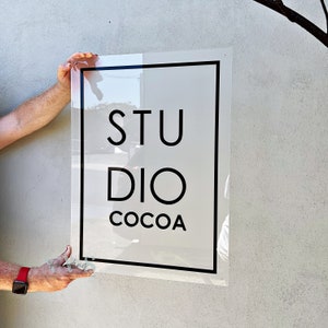 May include: A clear acrylic sign with the words "STUDIO COCOA" in black, centered on a white background, framed by a black border. The sign is held up against a light grey wall.