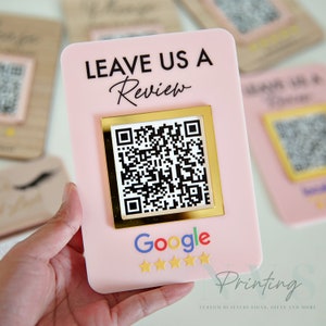 Puede incluir: Un letrero rosa con el texto "Leave Us A Review" en letras negras. Un marco dorado rodea un código QR en blanco y negro. Debajo del código QR está la palabra "Google" en letras de colores y cinco estrellas doradas. El texto "Printing" está en letras negras con el texto "Custom Business Signs, Gifts and More" en letras negras más pequeñas debajo.