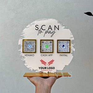 May include: A round, white acrylic sign with the text "SCAN to pay" in black. Three gold-framed QR codes are displayed below the text, labelled "VENMO", "CASH APP", and "PAYPAL". A red and white logo with the text "YOUR LOGO" is displayed below the QR codes.