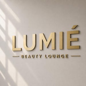 May include: A three-dimensional wall sign featuring the word LUMIÉ in large, brushed gold metallic letters, with the words BEAUTY LOUNGE in smaller gold letters underneath, flanked by two horizontal gold lines on a light, neutral-coloured wall.