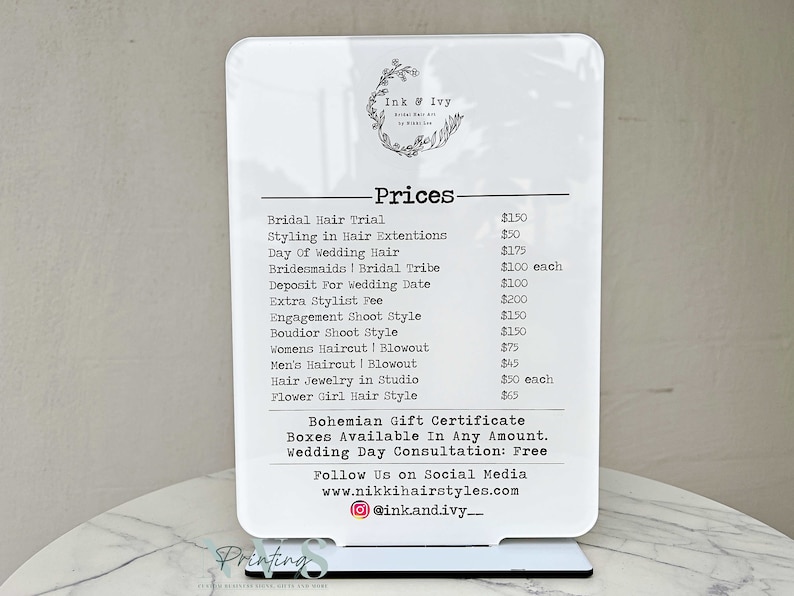 May include: White sign with black text listing prices for hair styling services. The sign reads "Ink & Ivy" and lists prices for services such as bridal hair trial, styling in hair extensions, day of wedding hair, bridesmaids, bridal tribe, deposit for wedding date, extra stylist fee, engagement shoot style, boudoir shoot style, women's haircut and blowout, men's haircut and blowout, hair jewelry in studio, and flower girl hair style. The sign also includes the text "Bohemian Gift Certificate Boxes Available In Any Amount. Wedding Day Consultation: Free" and "Follow Us on Social Media www.nikkihairstyles.com @ink.and.ivy_"
