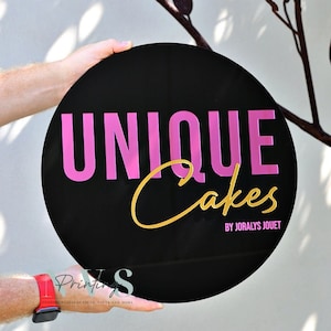 a round business signage with black background with UNIQUE in block letters on top in 3 hot pink mirror acrylic, CAKES under it in cursive font in 3D gold mirror acrylic and BY JORALYS JOUET on the bottom UV printed in hot pink colour
