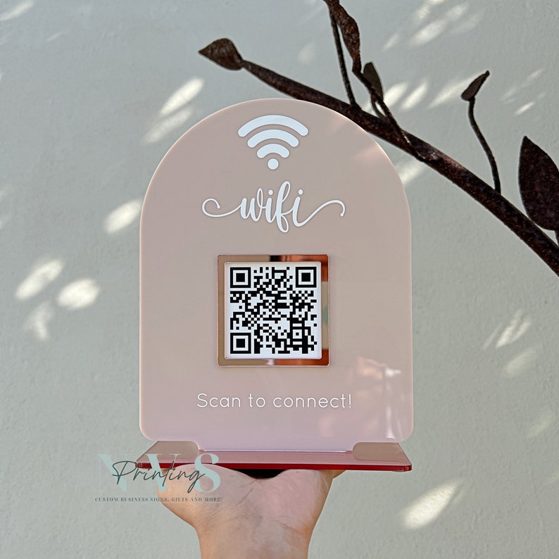 May include: A blush-coloured acrylic WiFi sign with a white WiFi symbol and script text. A QR code is centered below the text, with the words "Scan to connect!" below it. The sign is held up by a stand.