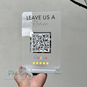Könnte beinhalten: Ein klares Acrylschild mit dem Text "LEAVE US A Review and receive $5 off of your next visit!"  Ein schwarz-weißer QR-Code wird unter dem Text angezeigt. Der Text "Google" befindet sich unter dem QR-Code mit fünf gelben Sternen.