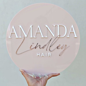 A round acrylic business sign in blush pink backing, with AMANDA in 3D gloss white acrylic in serif font, Lindley in cursive font and in 3D rose gold mirror, followed with the word HAIR on the bottom in 3D gloss white