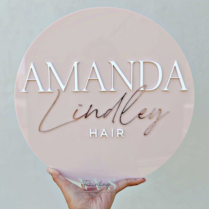 A round acrylic business sign in blush pink backing, with AMANDA in 3D gloss white acrylic in serif font, Lindley in cursive font and in 3D rose gold mirror, followed with the word HAIR on the bottom in 3D gloss white