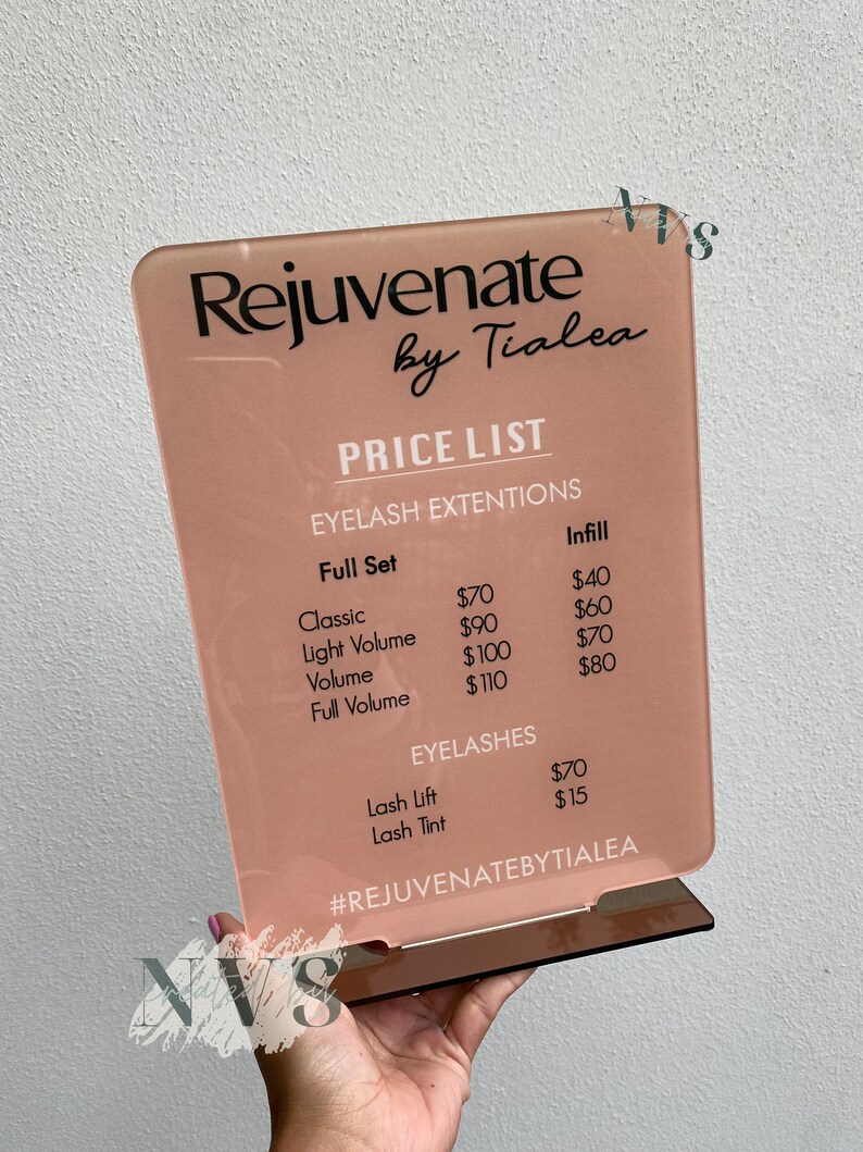 May include: A pink acrylic sign with the text "Rejuvenate by Tialea" and a price list for eyelash extensions and lash services. The prices are listed in US dollars. The sign has a hashtag at the bottom that reads "#REJUVENATEBYTIALEA".