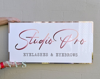 May include: A rectangular white sign with rose gold lettering and trim. The sign reads "Studio Pro" in a cursive font above the text "Eyelashes & Eyebrows" in a sans-serif font. The sign is held by a person.