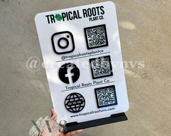 May include: White sign with black text and graphics. The sign reads "TROPICAL ROOTS PLANT CO." and includes social media icons and QR codes for Instagram, Facebook, and the company website.