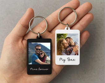May include: Two rectangular keychains with metal rings, each displaying a photograph. One is black with the text "Mine forever," the other is white with "My Bae." The keychains are held in a hand.