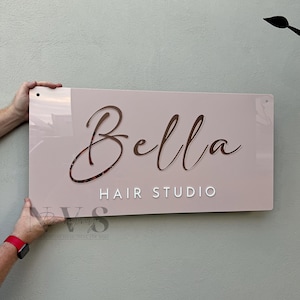 Könnte beinhalten: Ein rechteckiges Schild für einen Friseursalon. Das Schild ist hellrosa mit dem Namen "Bella" in Schreibschrift und den Worten "Hair Studio" in einer serifenlosen Schriftart. Das Schild wird von zwei Händen gehalten.