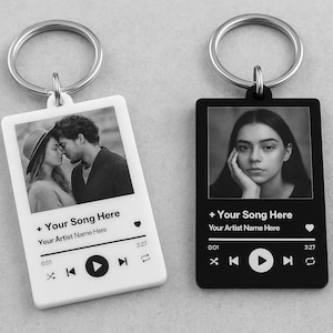 May include: Two rectangular keychains with silver key rings. One is white, the other black. Each features a black and white photo, music player controls, and text that reads "+ Your Song Here" and "Your Artist Name Here."