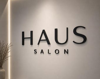 May include: A wall-mounted sign featuring the word HAUS in large, bold, black, three-dimensional lettering, with the word SALON positioned directly underneath in a smaller, matching black font, set against a smooth, light-colored wall surface.