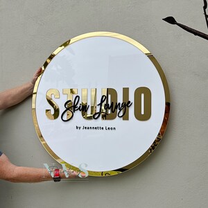 Acrylic Round Business Sign against the wall in white acrylic backing with gold mirror round border frame and 3D letters in gold mirror acrylic and black acrylic, skin lounge studio in 3D on the middle and business owner as business tagline