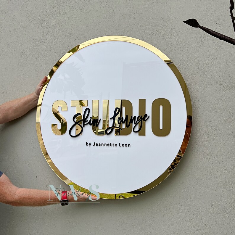 Acrylic Round Business Sign against the wall in white acrylic backing with gold mirror round border frame and 3D letters in gold mirror acrylic and black acrylic, skin lounge studio in 3D on the middle and business owner as business tagline