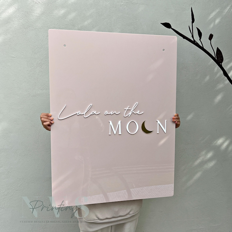Rectangle acrylic business sign in blush pink acrylic backing installed on the wall, Lola on the in 3D white acrylic in handwriting font, moon icon in 3D gold mirror, 2 mounting holes on top