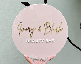 Könnte beinhalten: Ein rundes, rosafarbenes Acrylschild mit dem Text "Ivory & Blush Beauty Bar" in goldenen und weißen Buchstaben.