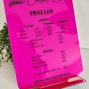 May include: Pink price list sign with black text for a lash and brow business called "Coastal Lashes". The sign lists prices for full sets of classic, hybrid, and volume lashes, as well as lash removal, lash tint, and lash lift and tint. It also lists prices for brow wax, brow tint, henna brow, and henna and wax. The sign also includes a price for a coastal bronze spray tan. The sign has the hashtag "#COASTALLASHESBYMILA" at the bottom.