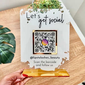 Lets get social sign with white splash background and gold mirror combination, QR code in the middle with Instagram logo, and instagram username under the QR code, and instruction to scan the Qr code to follow under the handle