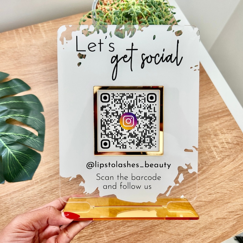 Lets get social sign with white splash background and gold mirror combination, QR code in the middle with Instagram logo, and instagram username under the QR code, and instruction to scan the Qr code to follow under the handle