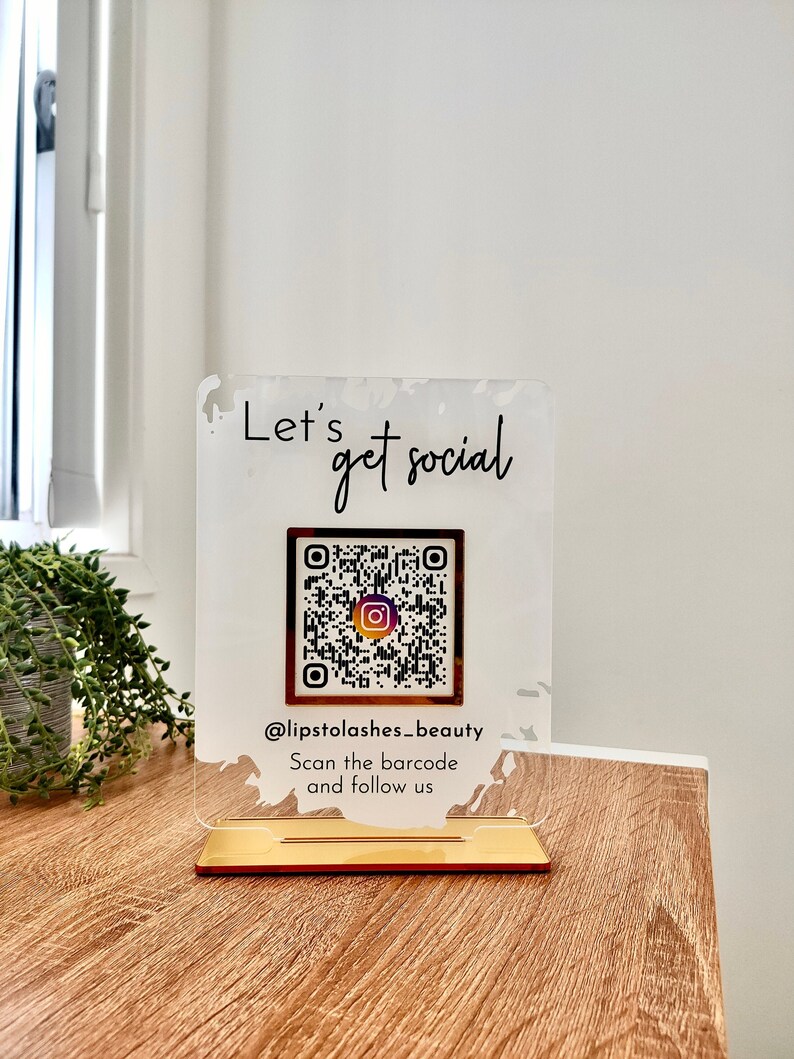 Lets get social sign with white splash background and gold mirror combination, QR code in the middle with Instagram logo, and instagram username under the QR code, and instruction to scan the Qr code to follow under the handle