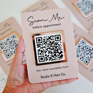 May include: A pink card with a gold frame around a black and white QR code. The text "Scan Me before appointment" is written in black cursive above the QR code. Below the QR code, the text "New client consultation form Studio K Hair Co." is written in black.