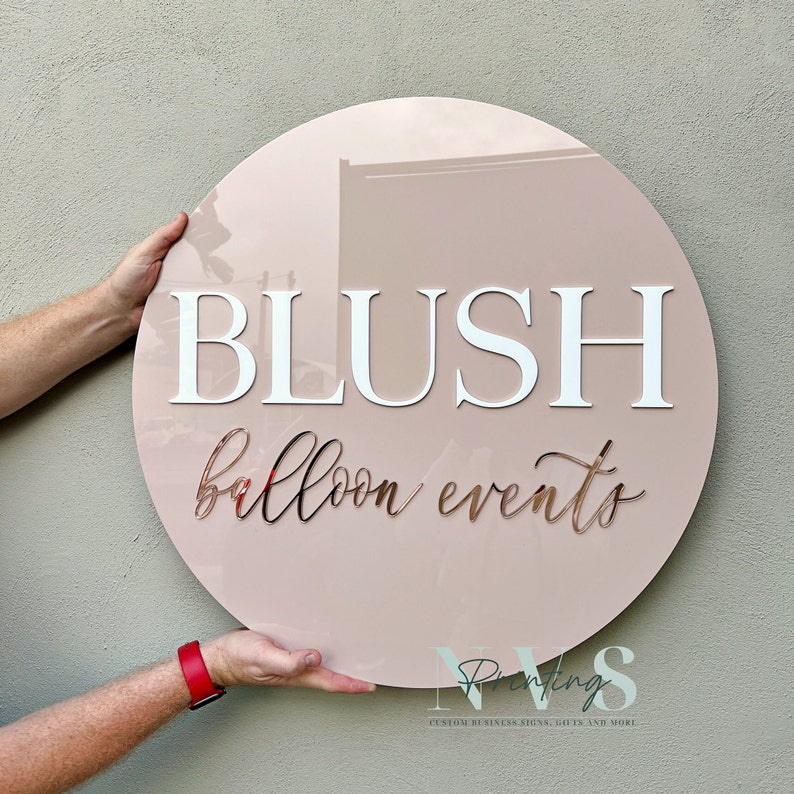 A round acrylic business sign in blush pink backing, BLUSH in capital in 3D gloss white acrylic in serif font, Balloon events in cursive font and in 3D rose gold mirror under it, it is hung on grey wall