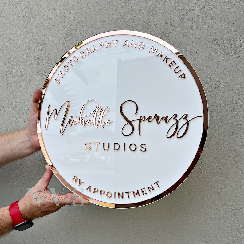 Acrylic Round Business Sign against the wall in white acrylic backing with rose gold mirror round border frame and 3D letters in rose gold mirror acrylic, photography and make up along the top and business studio name along the bottom