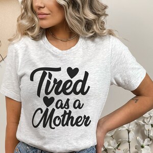Op de afbeelding: Een grijs T-shirt met de tekst "Tired as a Mother" in zwarte cursieve letters met drie zwarte harten.